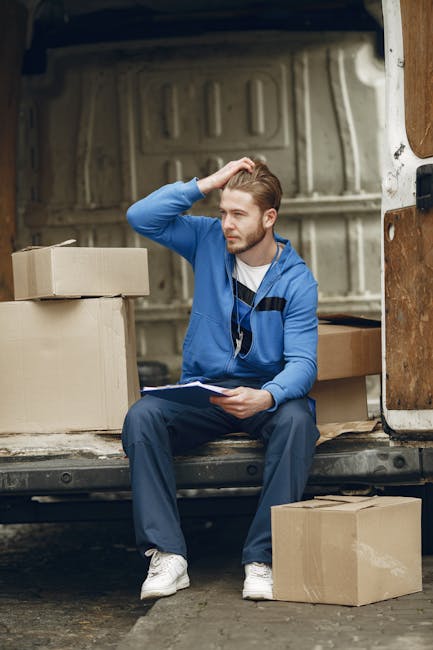 A young man with light brown hair and a beard sits on the edge of a moving van's open cargo area, surrounded by several cardboard boxes of various sizes, some sealed with packing tape. He is wearing a