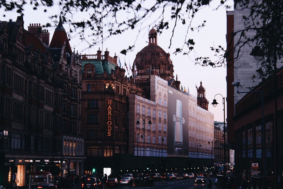 A street scene in Knightsbridge, featuring a large historic building with a domed roof, ornate architectural details, and a prominent Harrods sign illuminated on the facade. The building is partially obscured by a billboard and construction scaffolding, indicating active renovations. In the foreground, there are multi-story shops with large windows, street lamps, and parked cars along the curb. Tree branches with sparse leaves are visible at the top of the image, with ambient evening lighting casting a soft glow over the scene. This setting exemplifies the vibrant environment where home relocation and furniture transport services by Removal Company Knightsbridge might be involved in the process of packing and moving household items through the busy urban landscape near Harrods, SW1X.