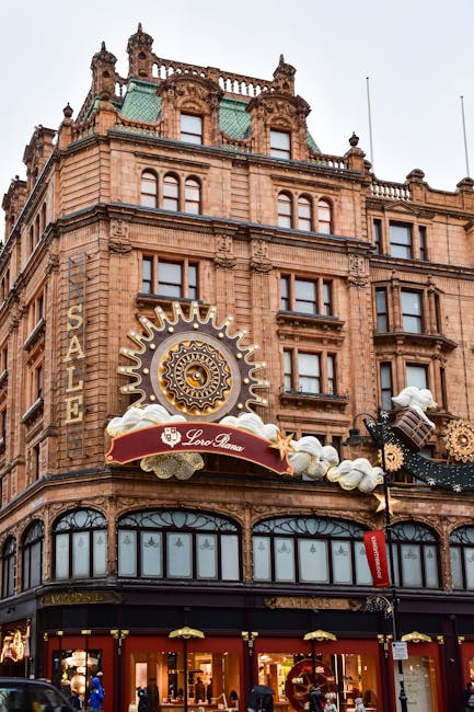 A street scene in Knightsbridge, featuring a large historic building with a domed roof, ornate architectural details, and a prominent Harrods sign illuminated on the facade. The building is partially obscured by a billboard and construction scaffolding, indicating active renovations. In the foreground, there are multi-story shops with large windows, street lamps, and parked cars along the curb. Tree branches with sparse leaves are visible at the top of the image, with ambient evening lighting casting a soft glow over the scene. This setting exemplifies the vibrant environment where home relocation and furniture transport services by Removal Company Knightsbridge might be involved in the process of packing and moving household items through the busy urban landscape near Harrods, SW1X.